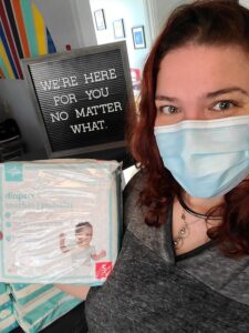 Image description: A photo of Center Manager Jess Marchbank wearing a surgical mask and holding a package of diapers, in front of a sign that says "We're here for you no matter what."