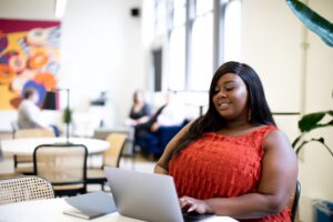 A Black woman in a bright orange dress smiles while looking at her laptop.