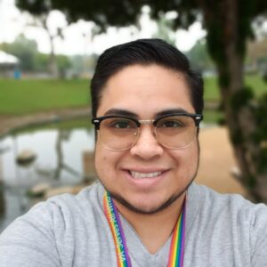 Image description: a photo of Jude Johnson, who wears glasses, a grey shirt, and a rainbow lanyard around his neck.