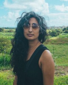 Image description: photo of Mohana Kute standing outside in front of a green field. Mohana wears sunglasses and a black shirt.